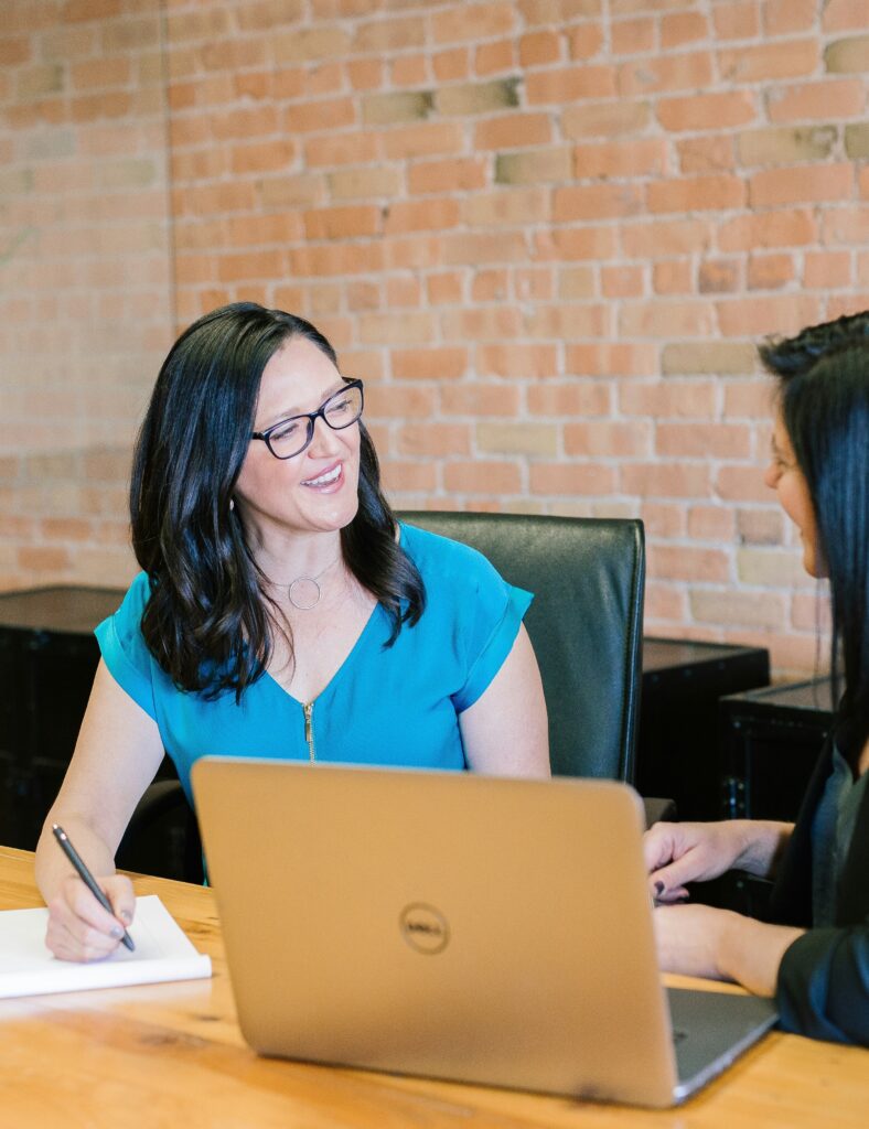Zwei Frauen lächeln und unterhalten sich am Schreibtisch mit Laptop und Notizblock vor einer Ziegelwand, während sie an digitalen Projekten und Weblösungen arbeiten.
