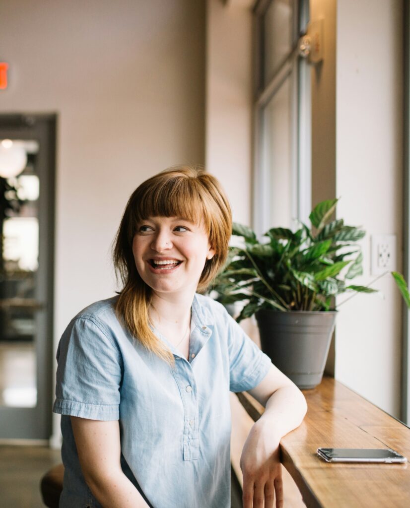 Eine lächelnde Person sitzt am Fenster neben einer Pflanze und einem Smartphone auf einem Holztisch, symbolisiert kreative Zusammenarbeit und digitale Lösungen für WordPress-Projekte in Bonn und dem Rhein-Sieg-Kreis.