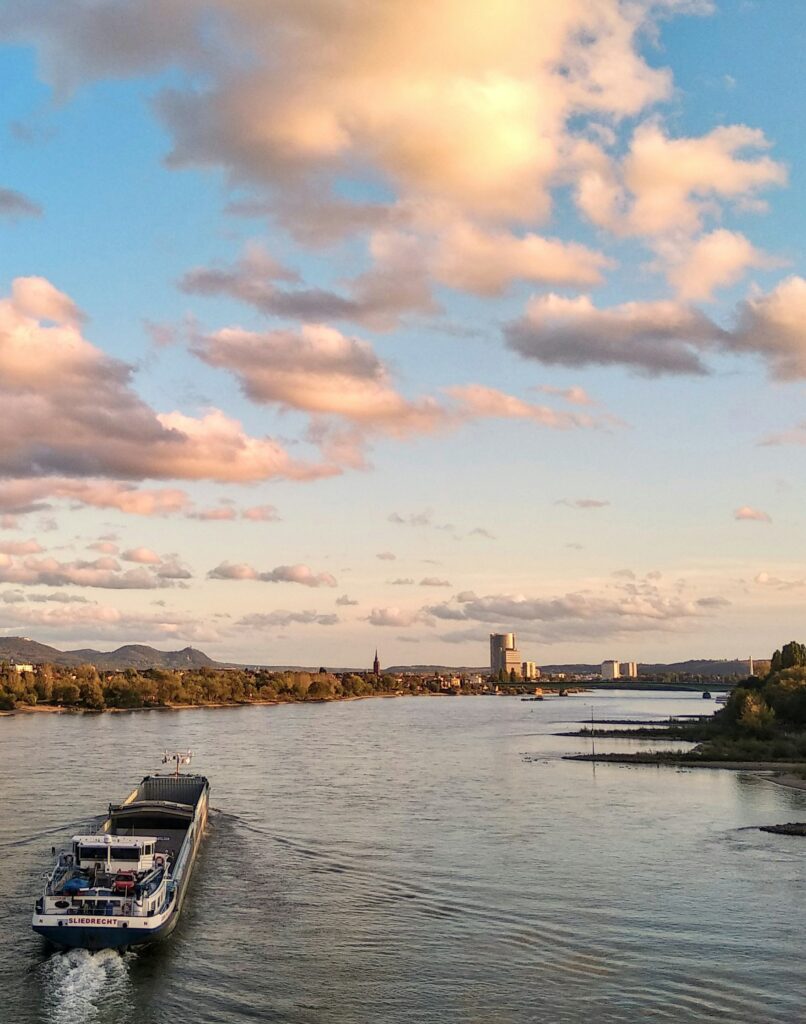 Frachtschiff auf breitem Fluss bei Sonnenuntergang mit grünen Ufern und bewölktem Himmel, symbolisiert innovative digitale Lösungen und erfolgreiche Webentwicklung, WordPress-Partner in Bonn und Rhein-Sieg-Kreis.